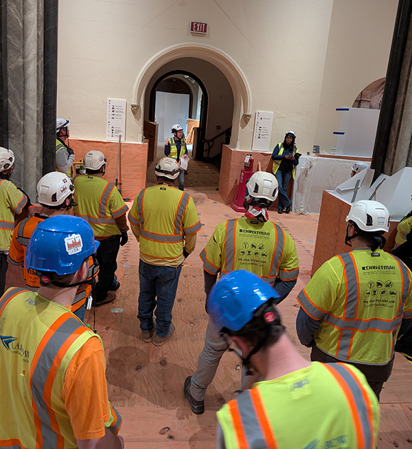 Group of people standing with hard hats and vests watching someone talk.