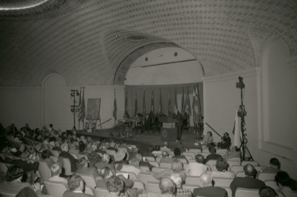 Image of the Baird Auditorium during a symposium looking towards the stage with audience members in seats. 