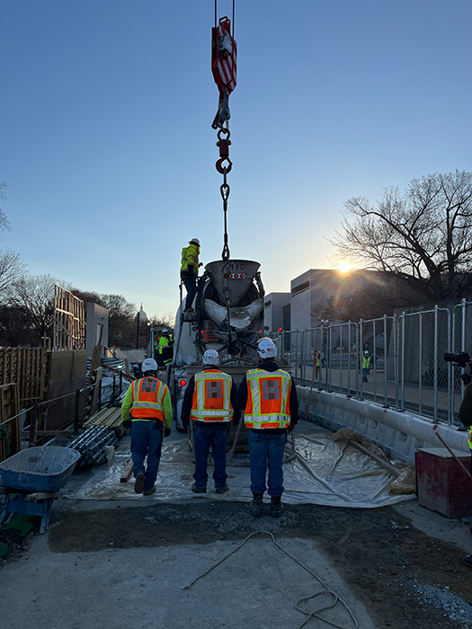 Image of people in safety vests and hard hats monitoring a truck delivery of concrete.